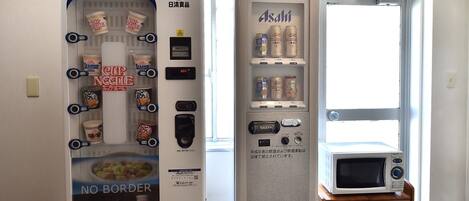 A vending machine with various food and drink options, a microwave, and a small table with a chair.