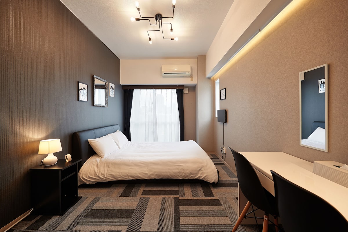 A hotel room with a bed, bedside table with a lamp, a desk with a chair, and a large mirror on the wall.