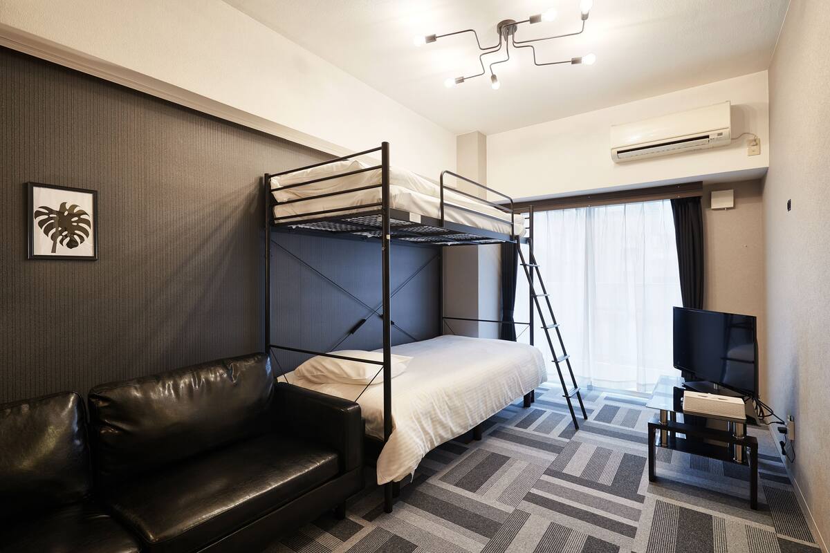 A hotel room with a bunk bed, a leather sofa, a television on a stand, and an air conditioning unit.