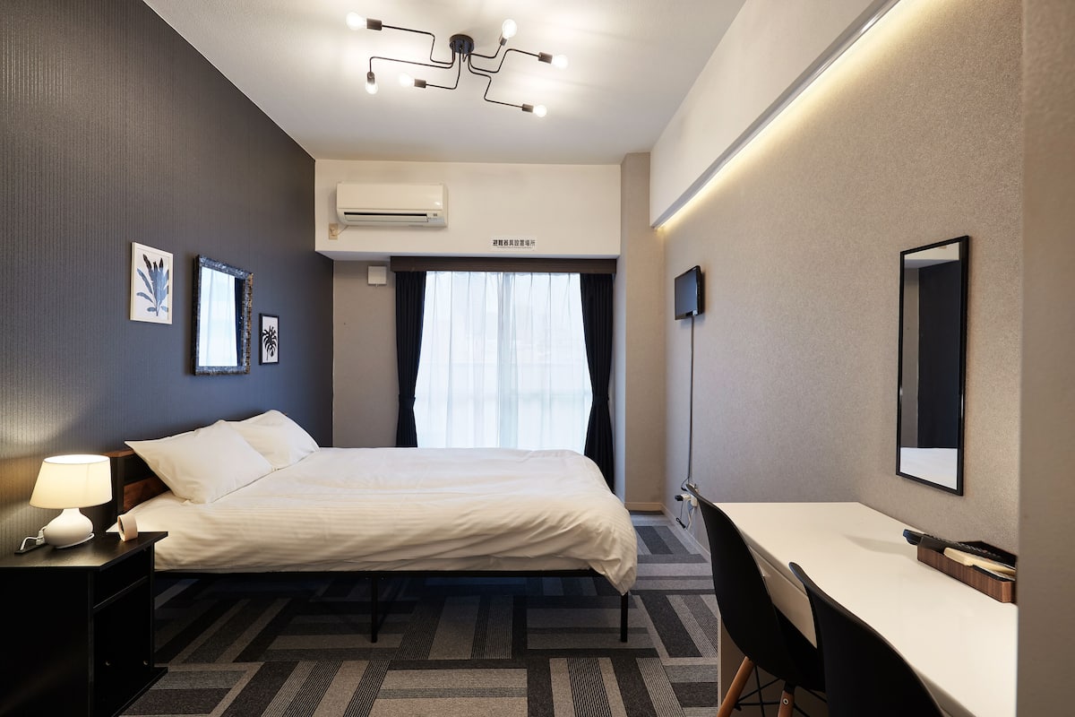 A hotel room with a bed, bedside table with a lamp, a desk, and a chair. There is an air conditioning unit on the wall and a flat-screen TV mounted on the wall.