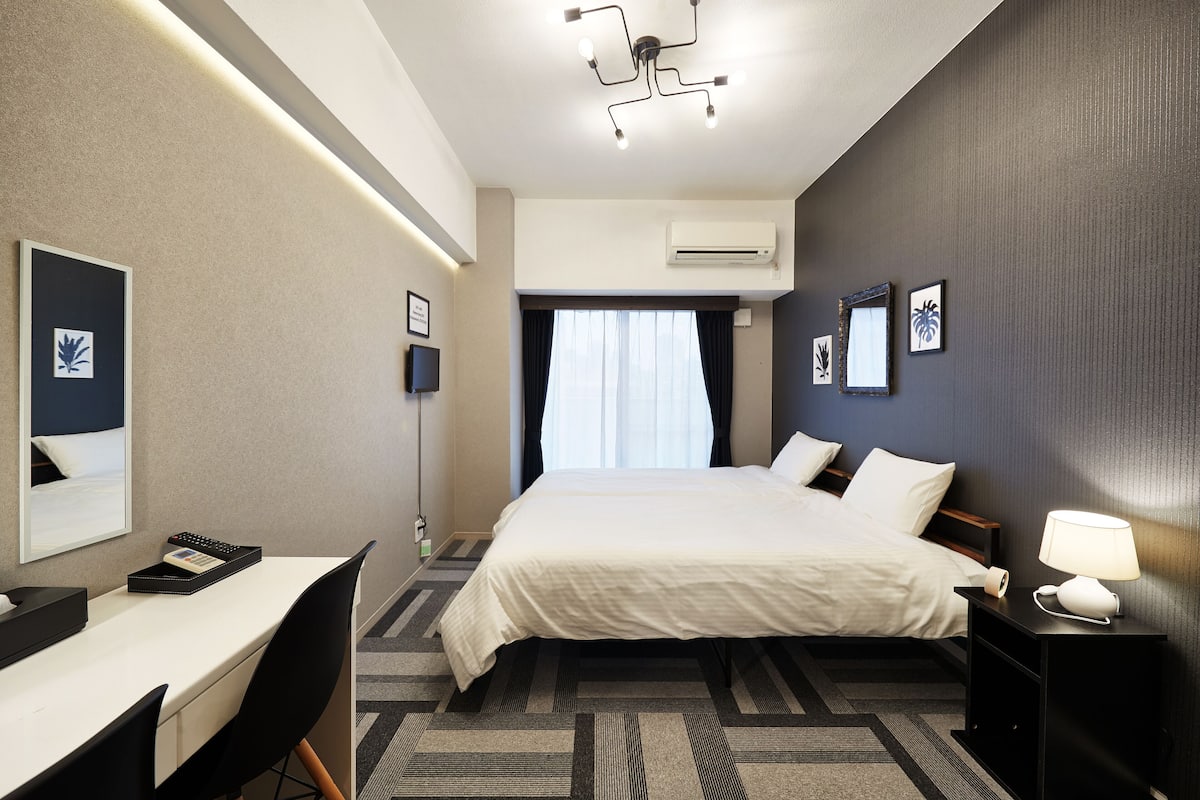 A hotel room with a bed, bedside table, desk, chair, and a ceiling light fixture.