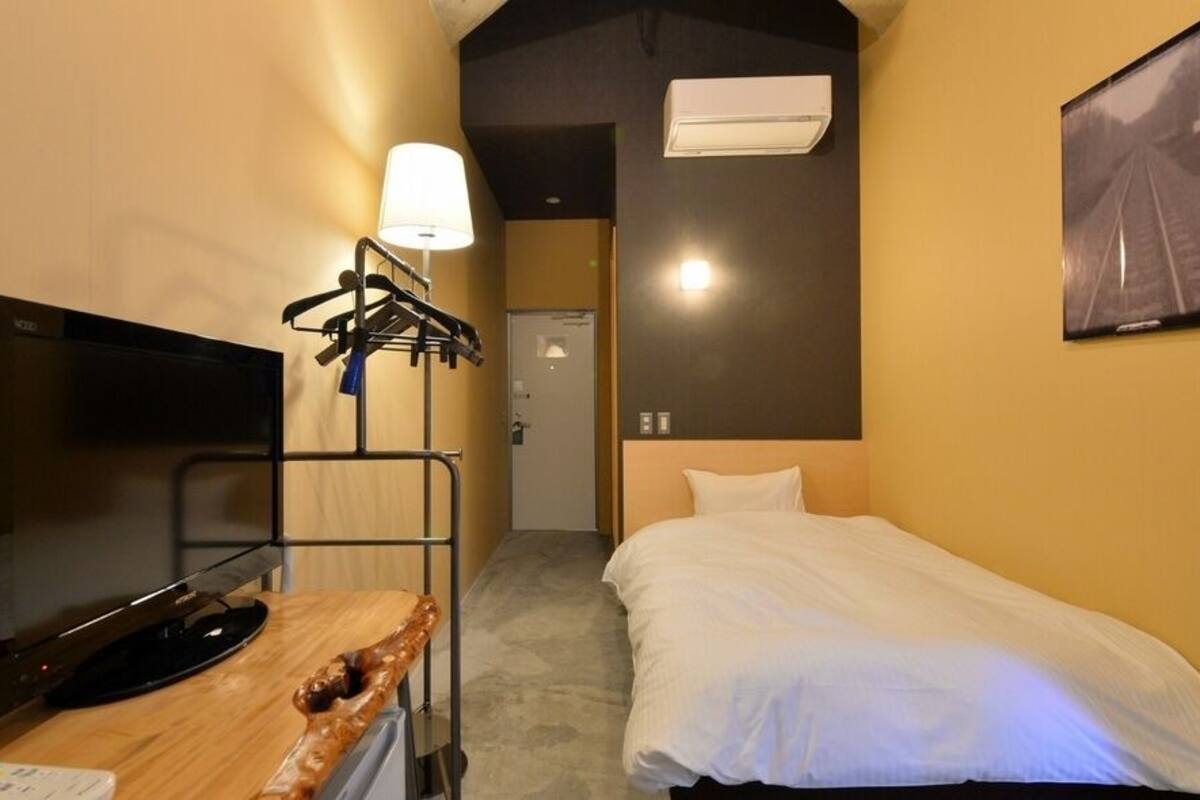 A hotel room with a bed, a TV, a desk with a lamp, a coat rack, and an air conditioner.