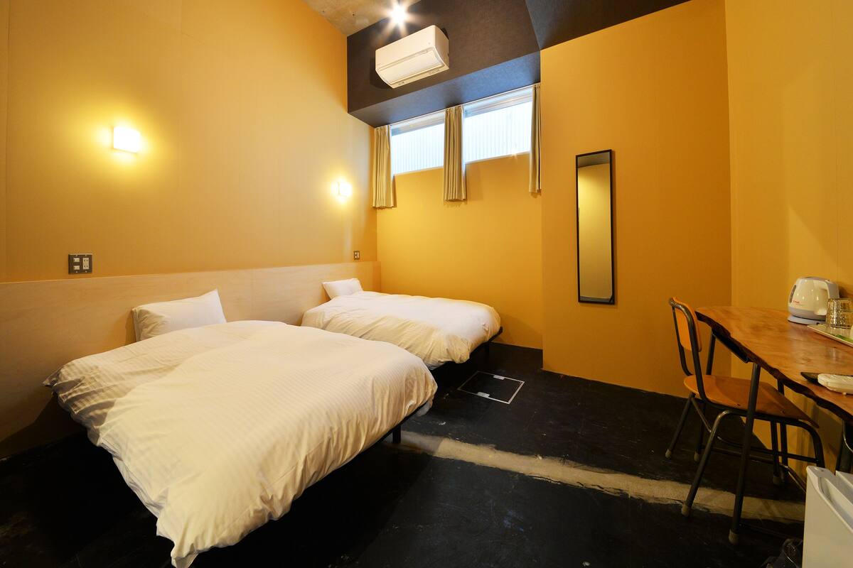 A hotel room with two beds, a desk, a chair, an air conditioning unit, and a window with curtains.