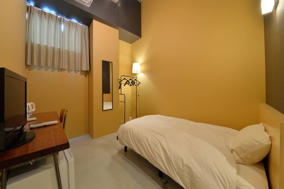 A hotel room with a bed, a desk with a computer, a chair, a bedside table, a lamp, and a window with curtains.