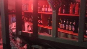 Un espace bar avec des étagères remplies de bouteilles, une personne debout près du comptoir et des caisses de bouteilles posées sur le sol.