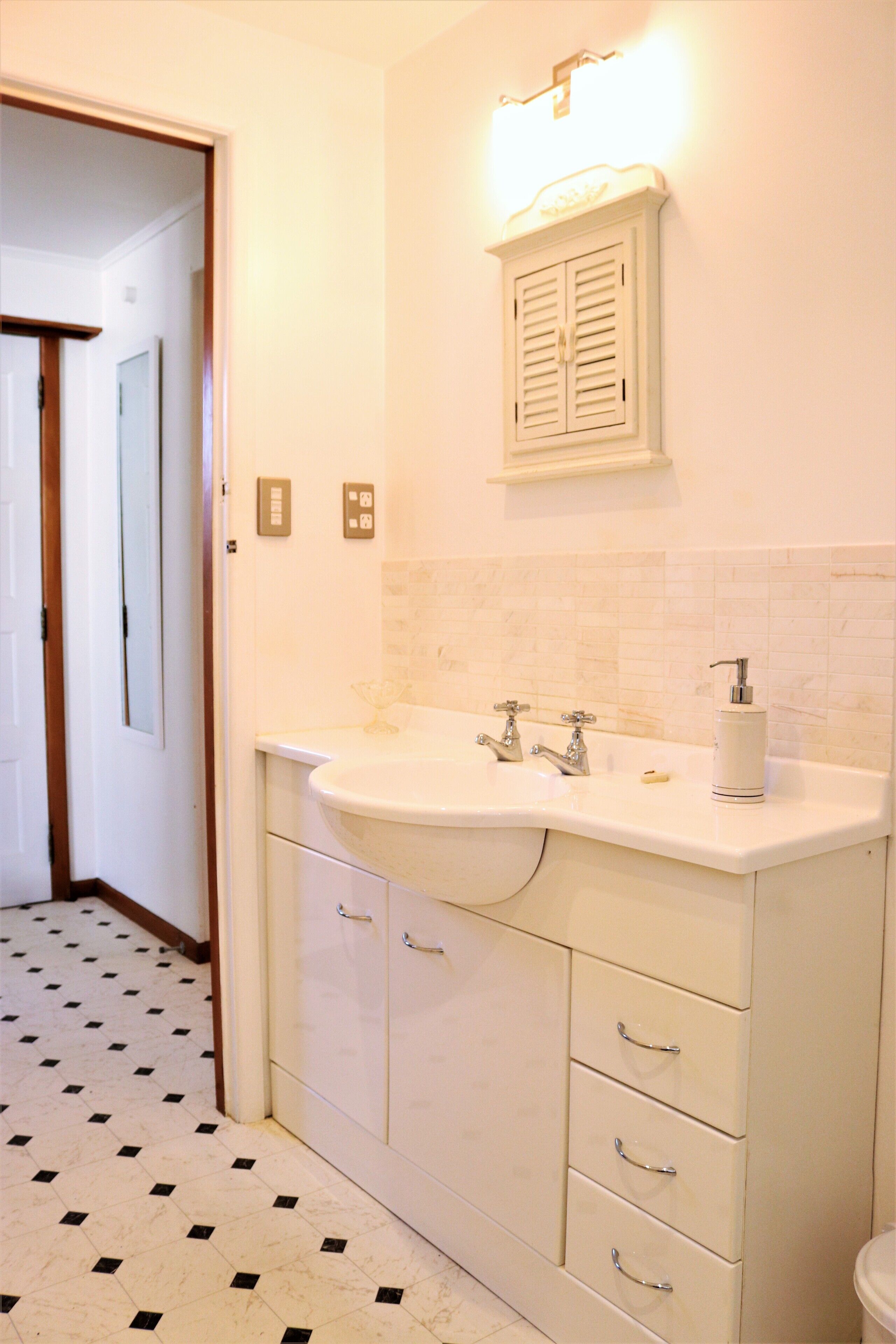 A bathroom with a white sink, two faucets, a soap dispenser, and a mirror above the sink. There are three drawers below the sink and a toilet to the right. The floor has a black and white tile pattern.