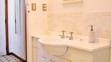 A bathroom with a white sink, two faucets, a soap dispenser, and a mirror above the sink. There are three drawers below the sink and a toilet to the right. The floor has a black and white tile pattern.