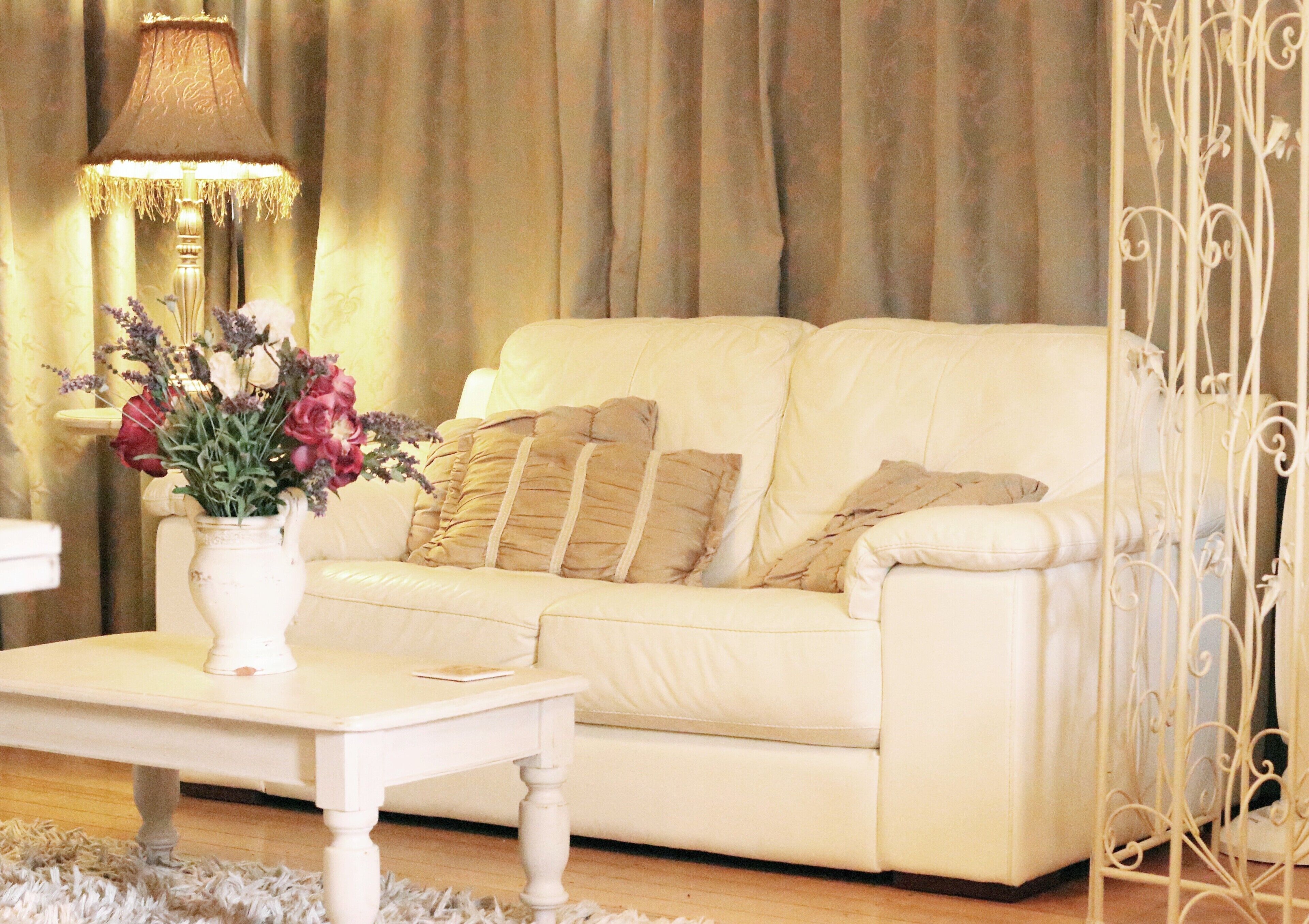 Un sofá de cuero blanco con varios cojines, una mesita auxiliar con un florero y una lámpara decorativa.