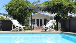 Una piscina con dos sillas reclinables blancas, un porche cubierto y una casa con techo azul al fondo.