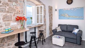 A cozy room with a stone wall, a wooden table with stools, a gray sofa, and a large window overlooking the sea.