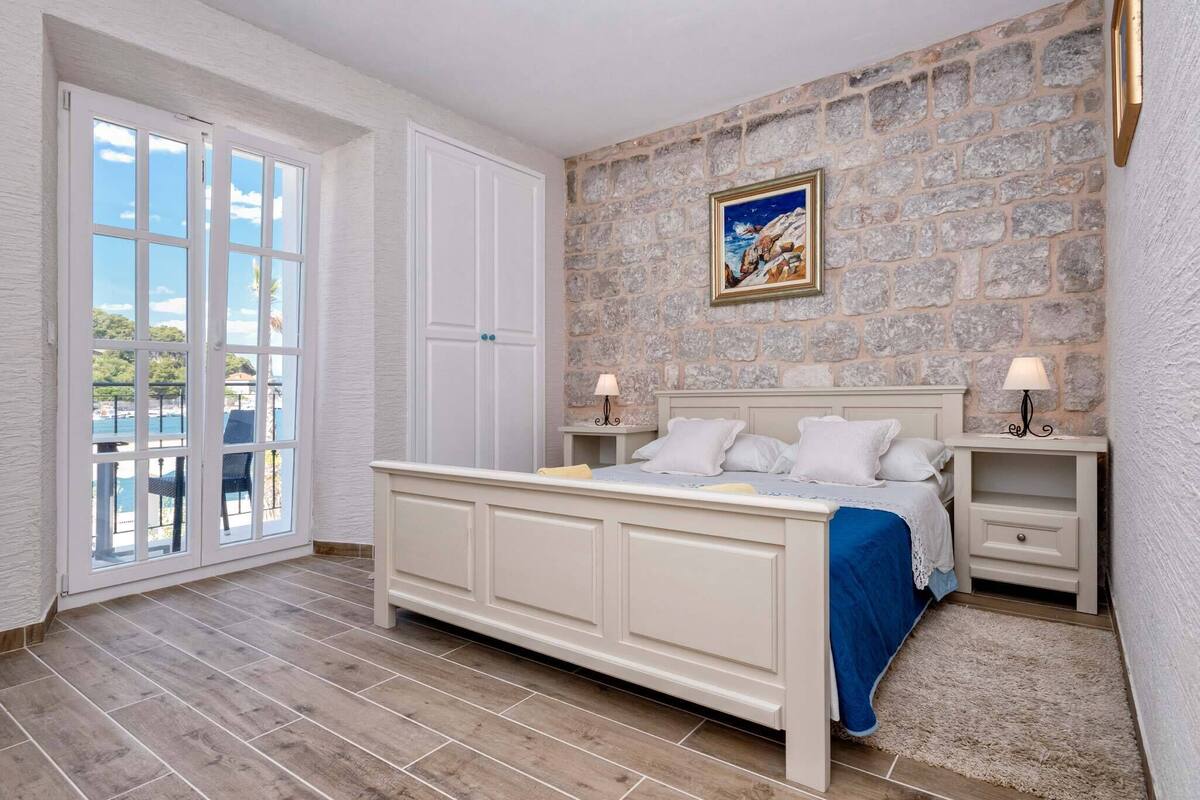 A bedroom with a stone wall, a bed with white bedding, two bedside tables with lamps, and a view through a glass door.