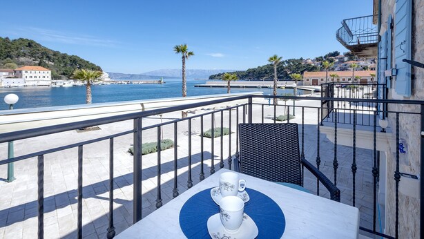 A balcony with a table set for two, overlooking a coastal area with a clear view of the sea and a hilly landscape in the background.