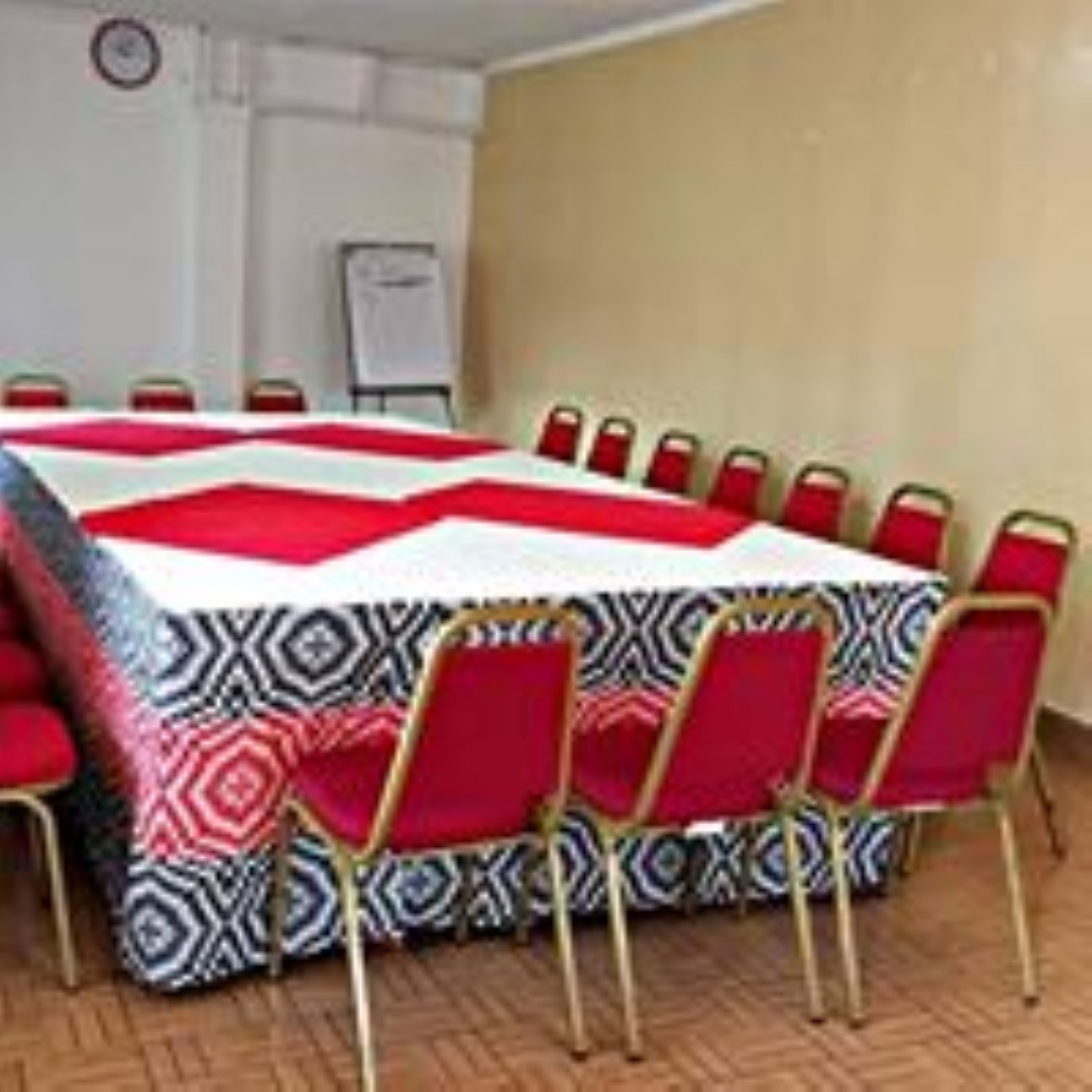 Un salón de conferencias con una gran mesa cubierta por un mantel estampado, rodeado de sillas rojas con patas doradas.