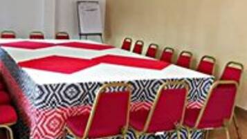 Un salón de conferencias con una gran mesa cubierta por un mantel estampado, rodeado de sillas rojas con patas doradas.