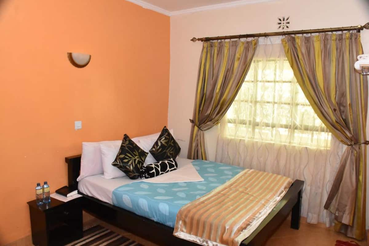 A bedroom with a bed, pillows, a bedside table, a window with curtains, and a wall-mounted light fixture.