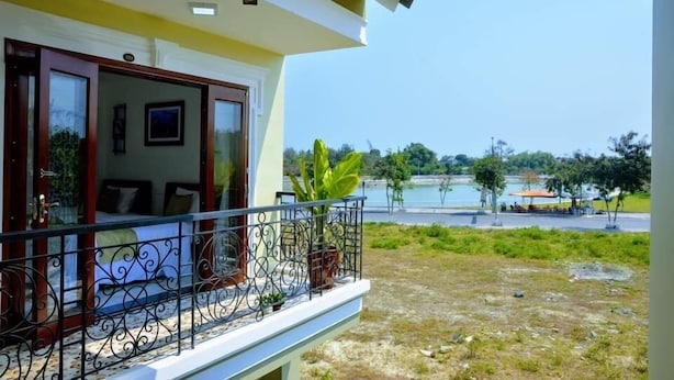A balcony with a metal railing, a bed with a white headboard, and a view of a body of water and greenery in the background. - Hoi An Lake Shore Homestay (Da Nang)
