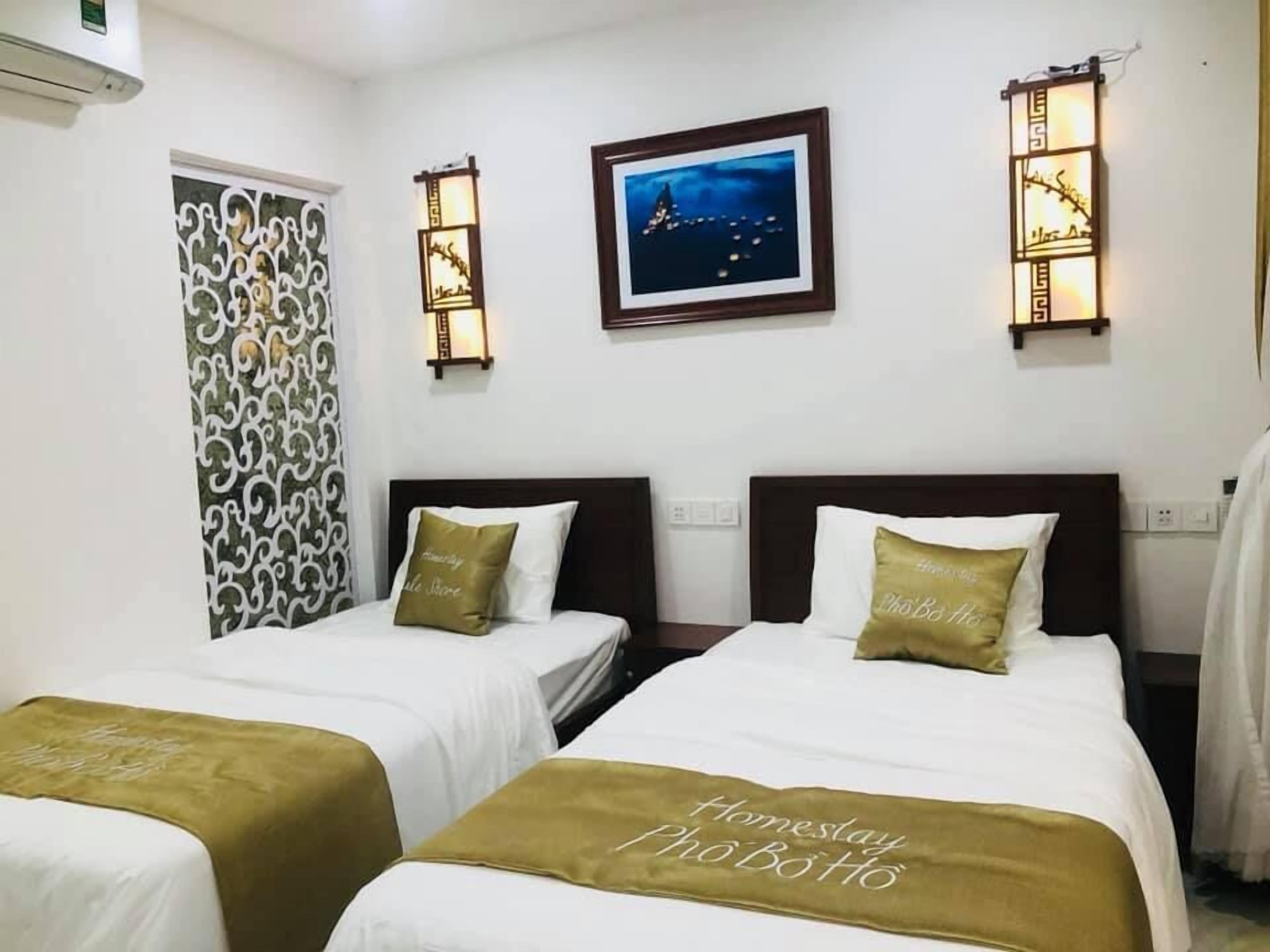 A hotel room with two beds, each with a white bedspread and a gold accent pillow. There is a framed picture on the wall above the beds, and two wall-mounted lamps.