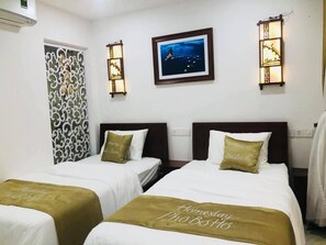 A hotel room with two beds, each with a white bedspread and a gold accent pillow. There is a framed picture on the wall above the beds, and two wall-mounted lamps.