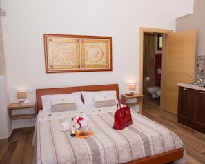 A neatly made bed with a striped bedspread, a red handbag, and a bottle of champagne on the pillows. There is a wooden headboard, two bedside tables with lamps, and a wall-mounted artwork above the bed. - La Dimora di Marco (Melendugno)