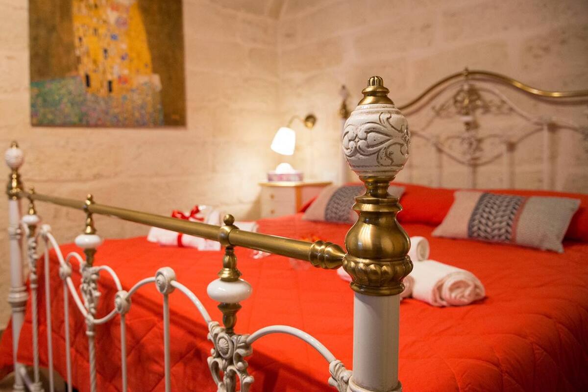 Un lit avec une couverture rouge, une tête de lit en laiton et un tableau accroché au mur.