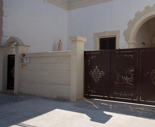 A gated entrance with a decorative metal gate, a white wall, and a door with a number plate. - La Dimora di Marco (Melendugno)