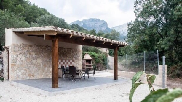 Eine Pergola aus Stein und Holz mit Tisch und Stühlen, vor einer Berglandschaft.