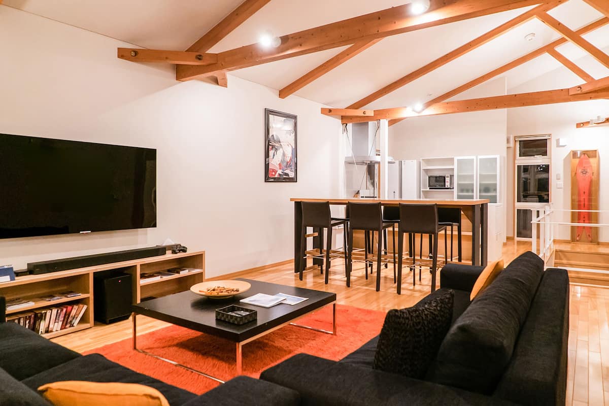A modern living room with a large flat-screen TV, a black sofa, a wooden dining table with chairs, and a kitchen area in the background.