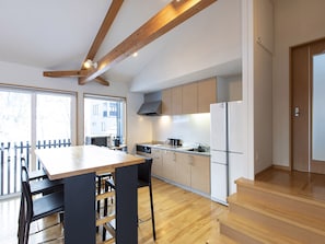 A modern kitchen with wooden beams, a dining area with a table and chairs, and a view of the outside through large windows. - Casi 67 Chalet (Kutchan)