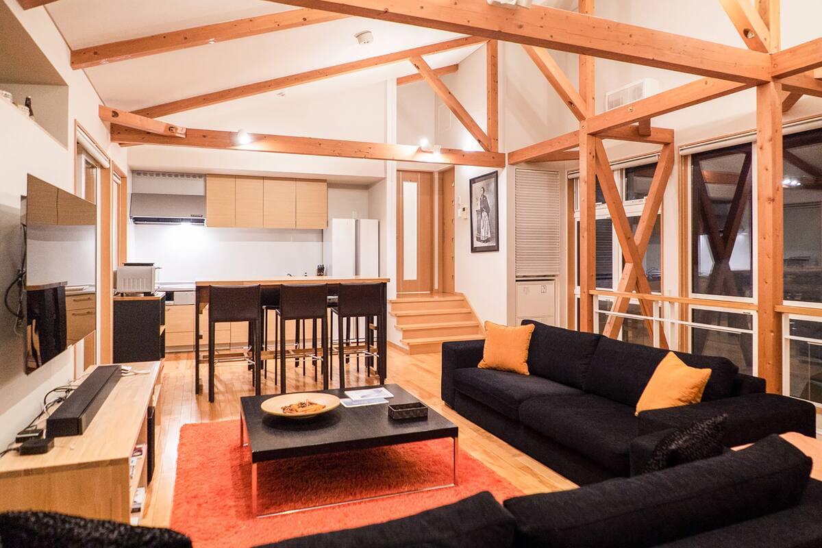 A modern living room with a black sofa, wooden beams, a dining area with chairs, and a kitchen in the background.
