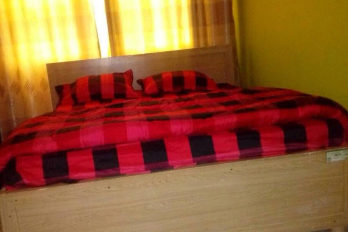 Una cama con manta a cuadros, cabecero de madera y ventana con cortinas.
