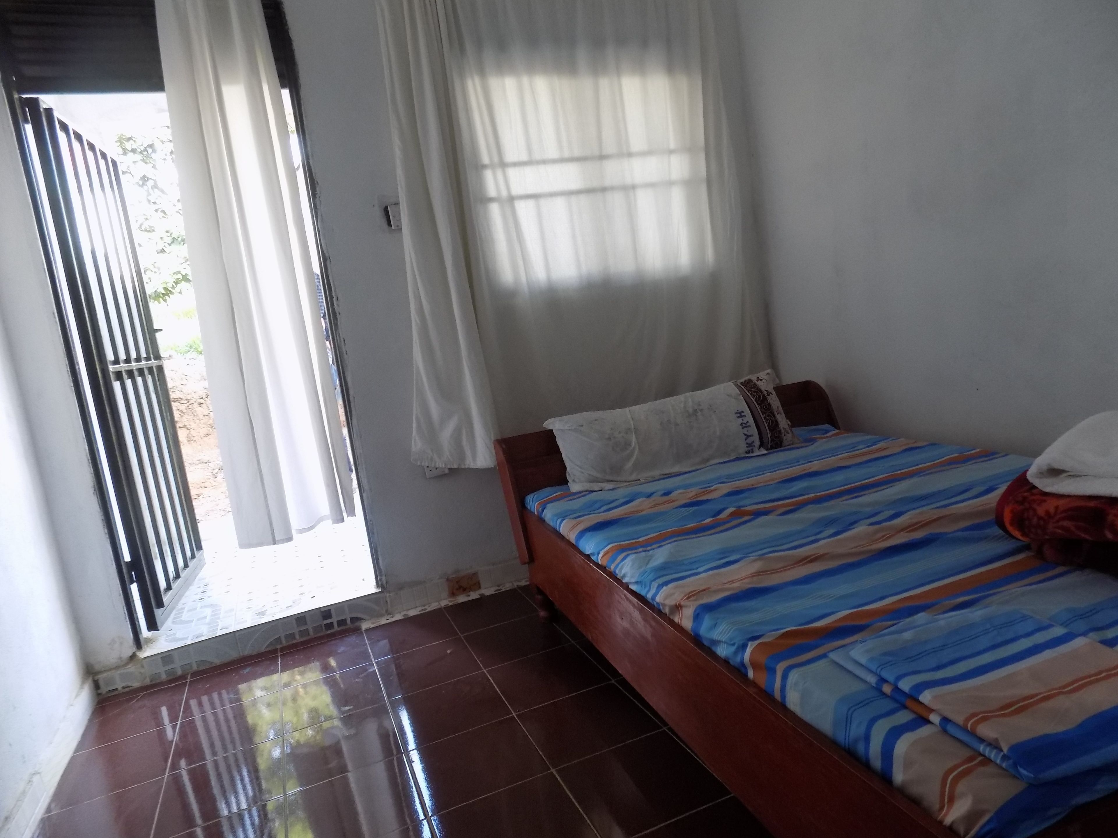 Una habitación con cama, ventana con cortinas, puerta que da al exterior y piso de baldosas.