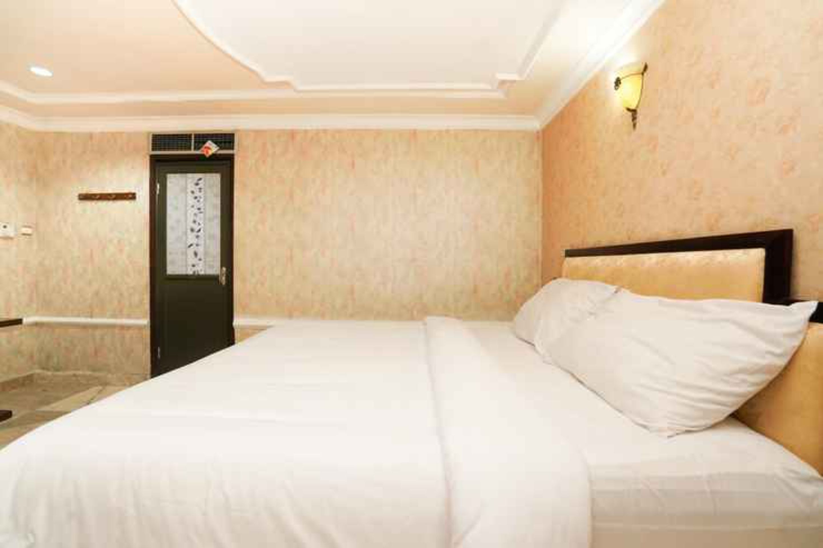A hotel room with a large bed, a door with a decorative panel, and a wall-mounted light fixture.