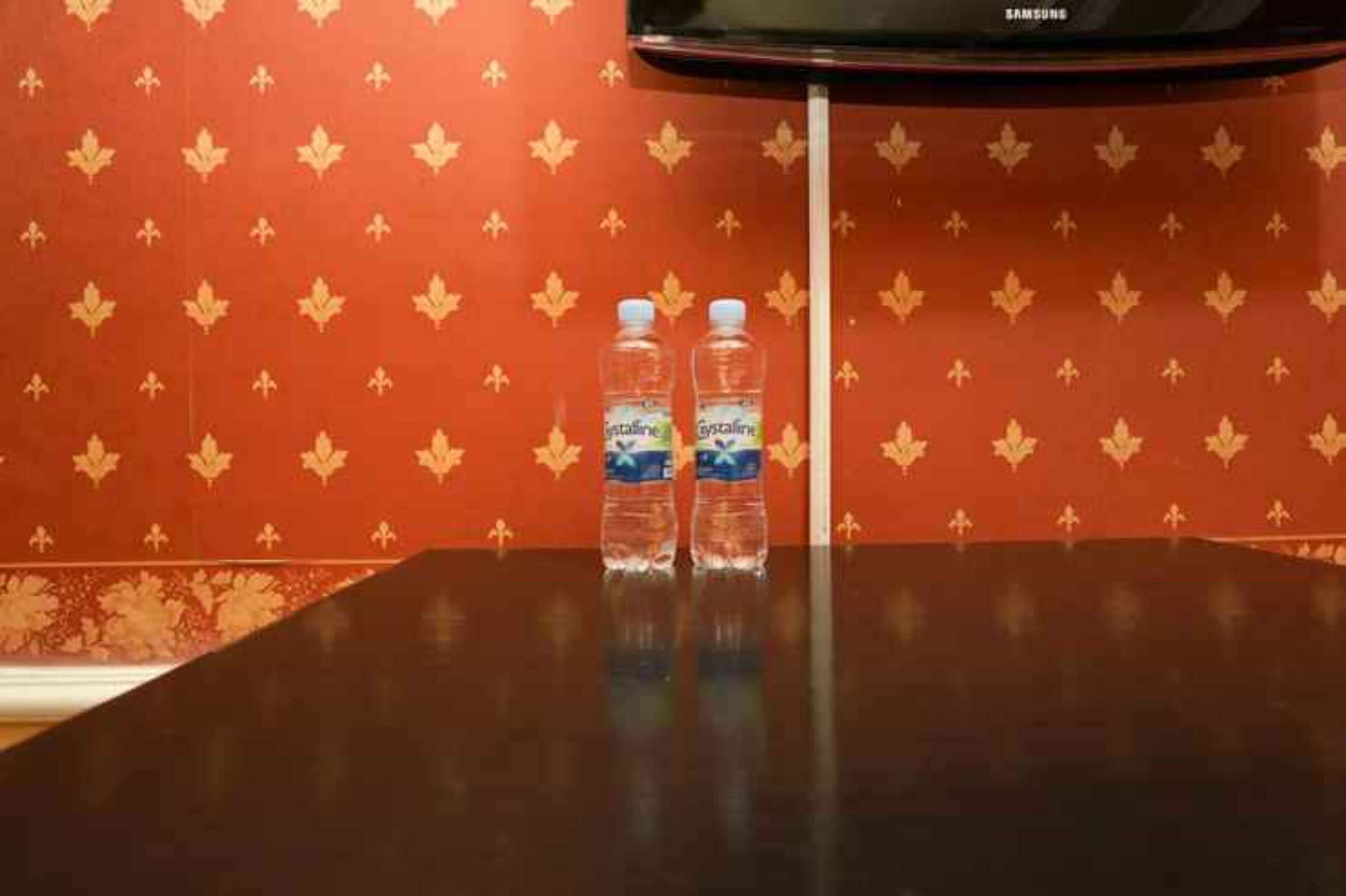 A room with a red patterned wallpaper, a Samsung TV mounted on the wall, and two water bottles on a table.