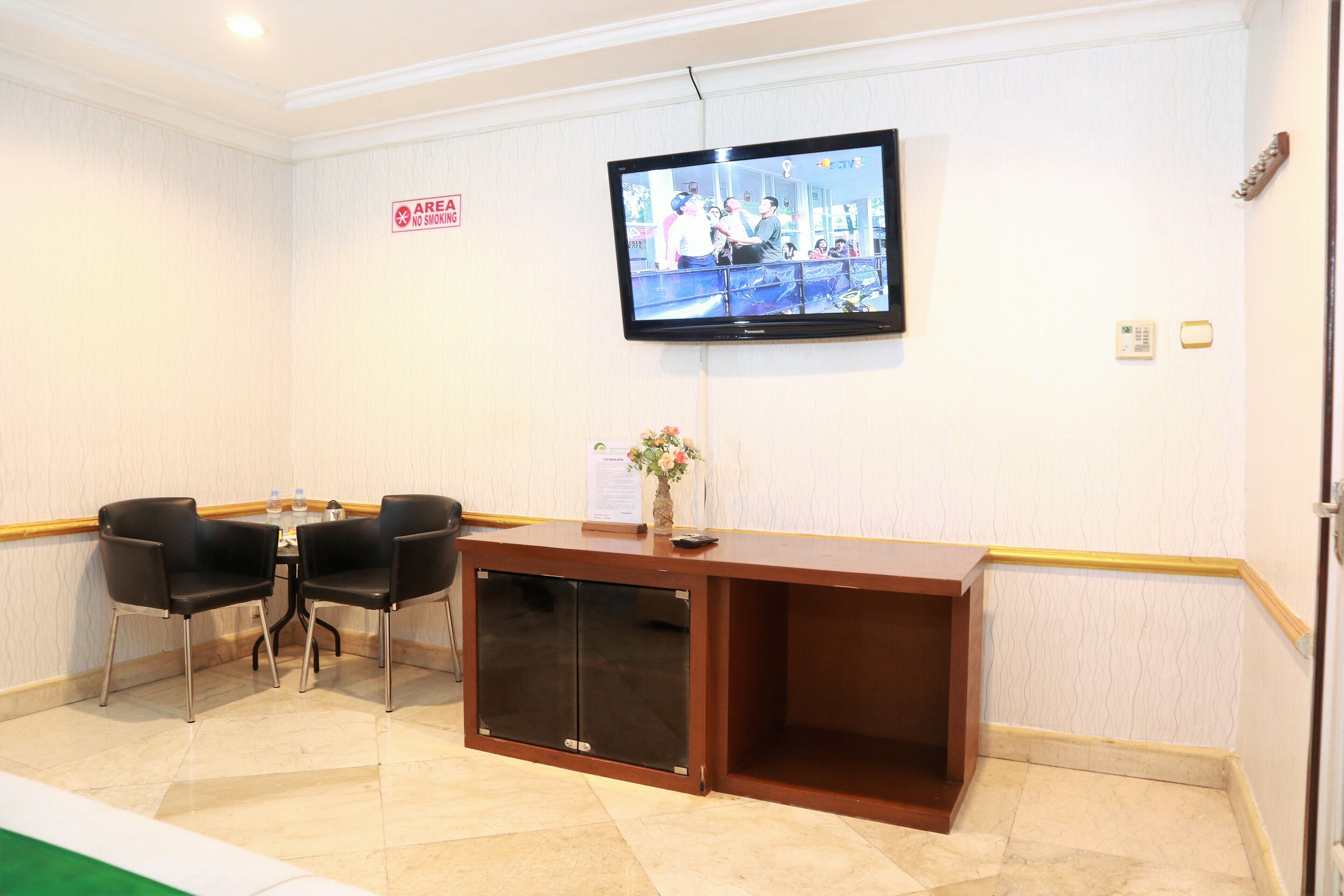 Zona de recepción de un hotel con una televisión montada en la pared, un mostrador de recepción de madera, dos sillas negras, una pequeña mesa con un jarrón de flores y un letrero de 'No Fumar'.