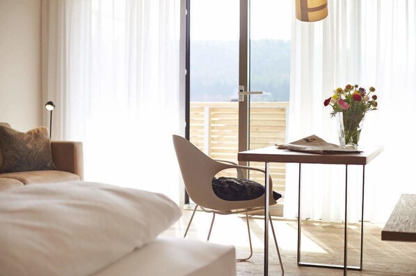 Un dormitorio con cama, una silla, una mesa con un florero y vistas al exterior a través de un amplio ventanal.