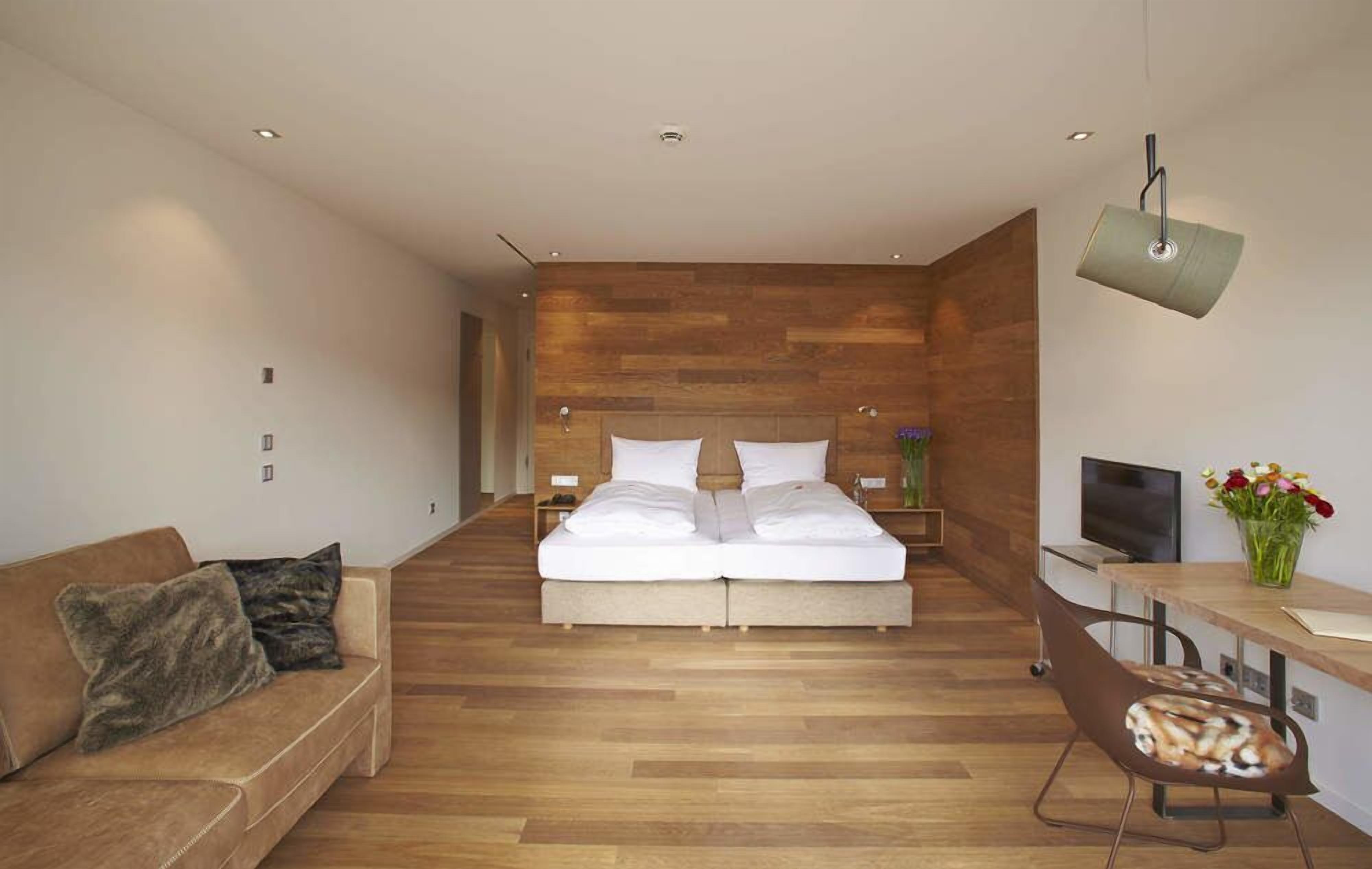 A modern bedroom with a wooden headboard, a double bed, a sofa, a desk with a TV, and a vase of flowers.