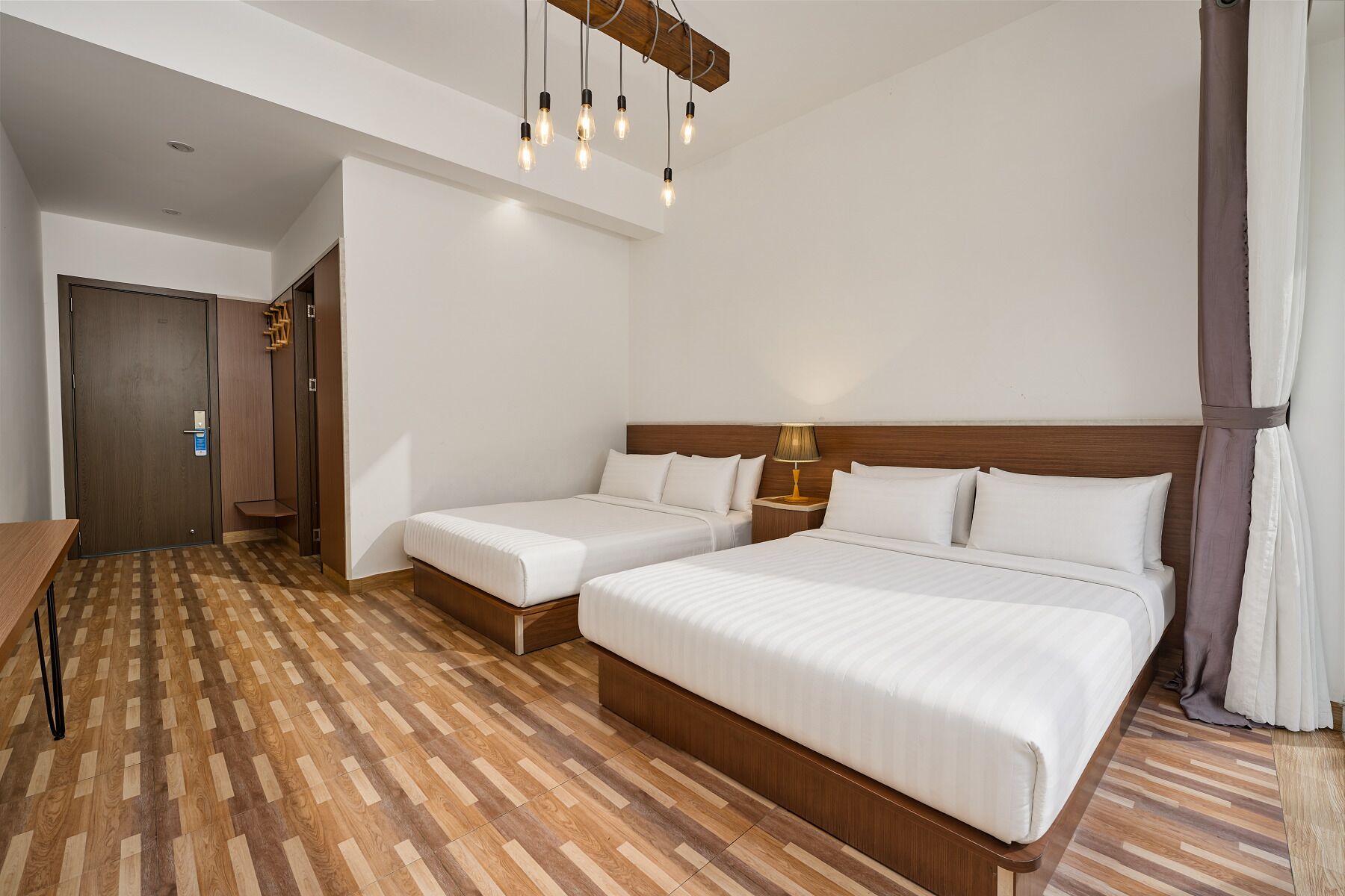 Habitación de hotel con dos camas, piso de madera, cabecero de madera, mesita de noche con lámpara y una puerta con manija.