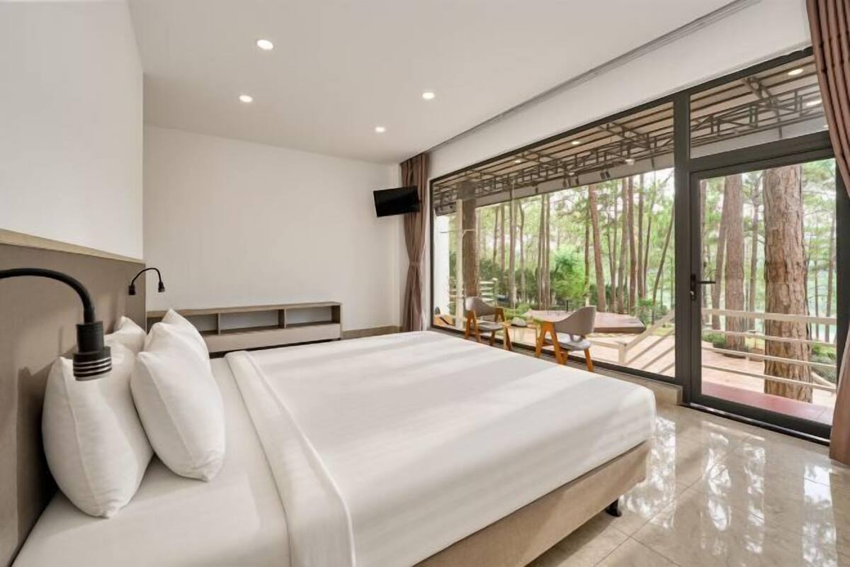 A modern bedroom with a large bed, a TV, and a view of a forest through a glass door.