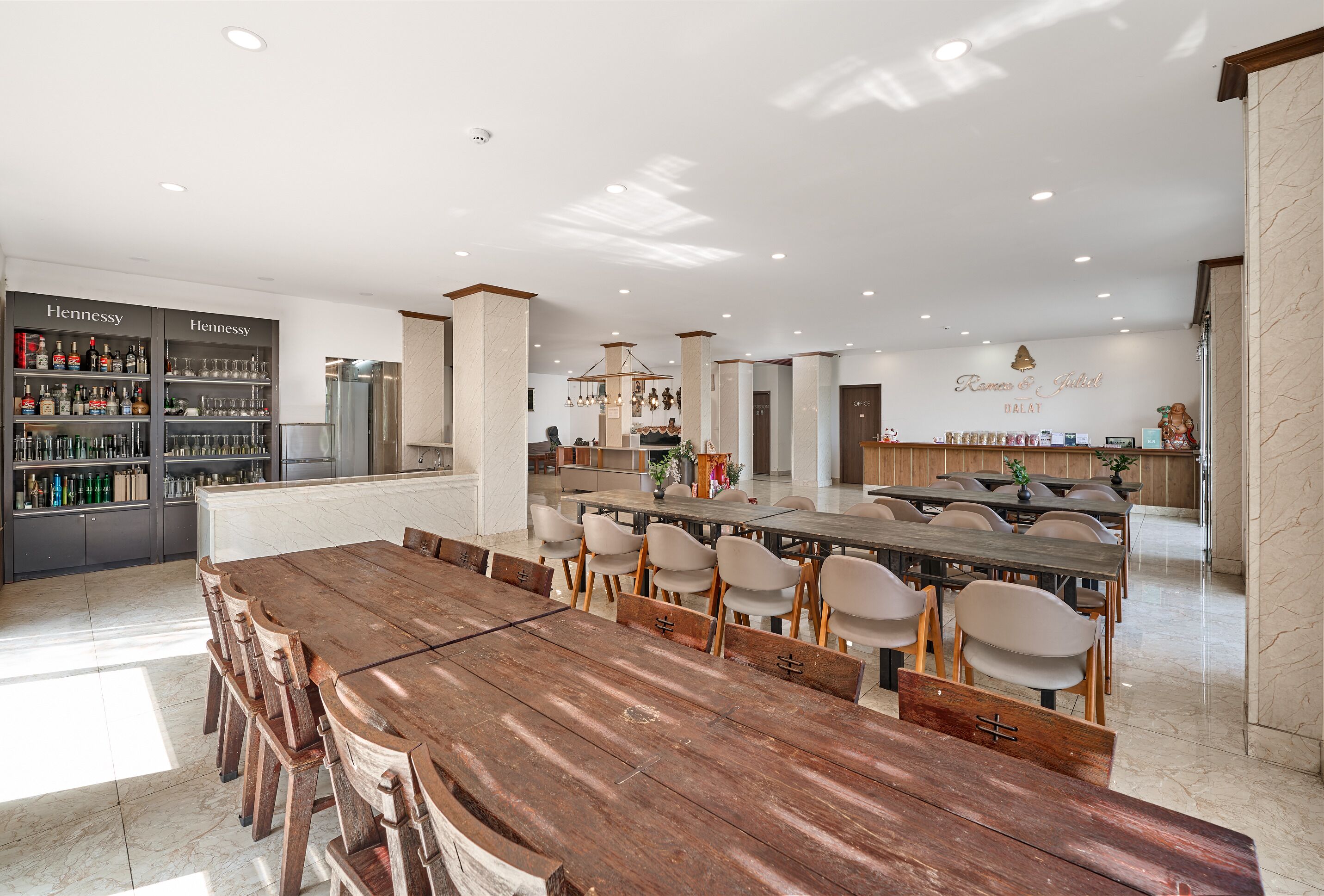 A modern dining area with a long wooden table, chairs, a bar with liquor bottles, and a sign for 'Ramos & Galat HALAT.'