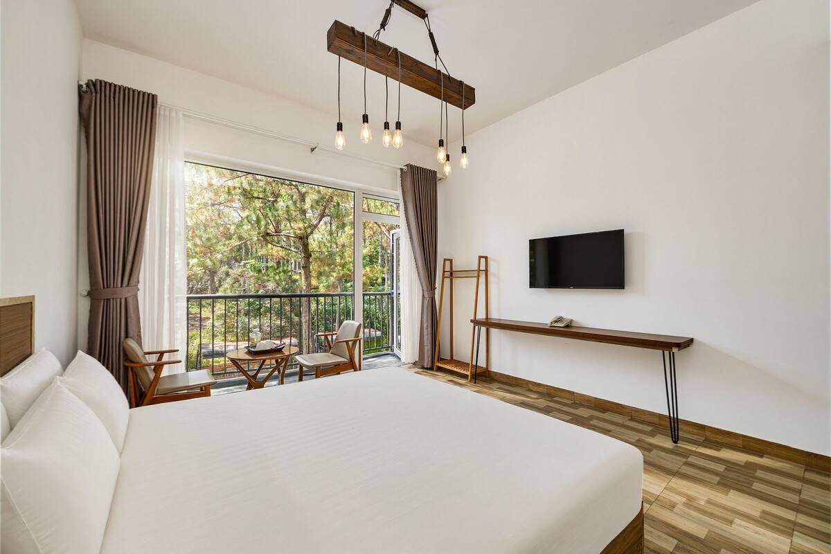 A hotel room with a large bed, a desk, a TV, and a balcony with outdoor seating.