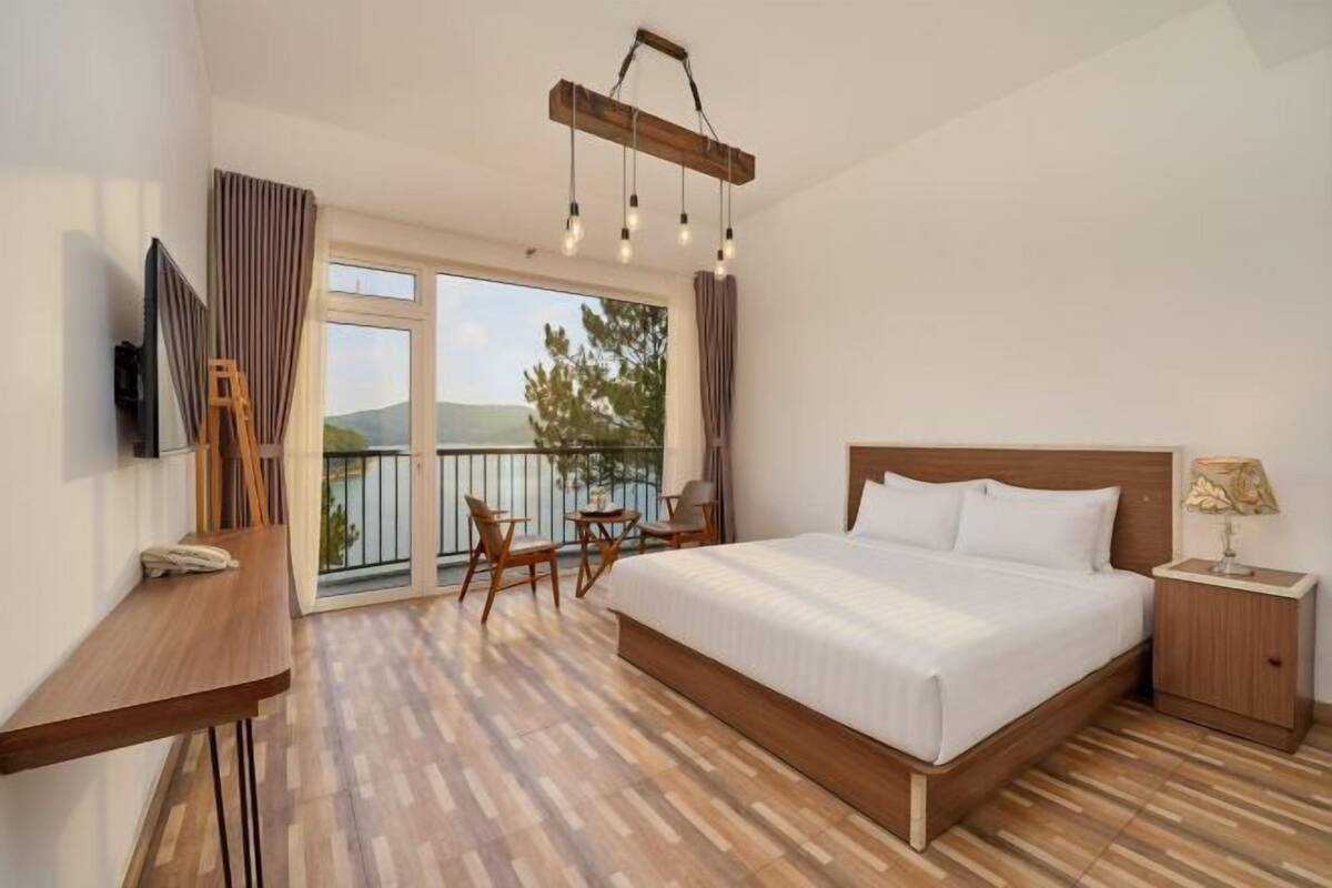 A bedroom with a large bed, a desk, a chair, a TV, a nightstand, and a balcony with a view.