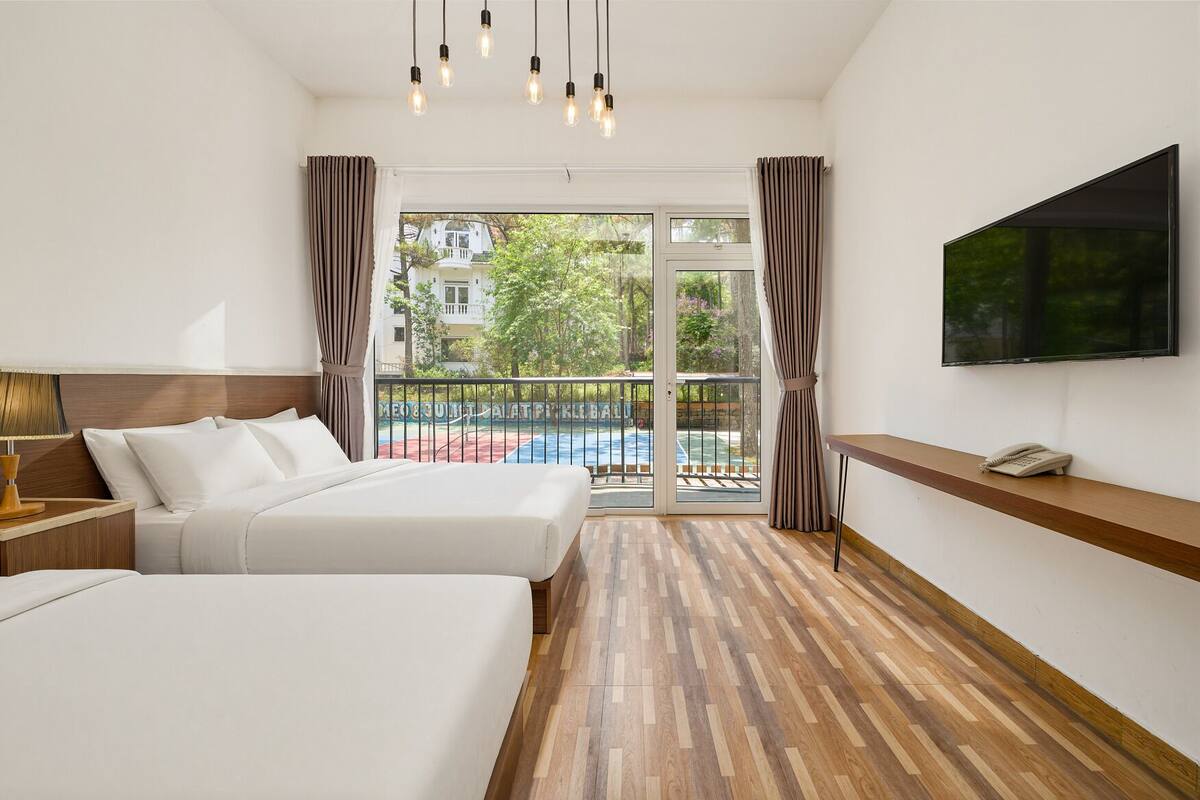 A hotel room with two beds, a TV, a phone, and a balcony with a view of a tennis court.