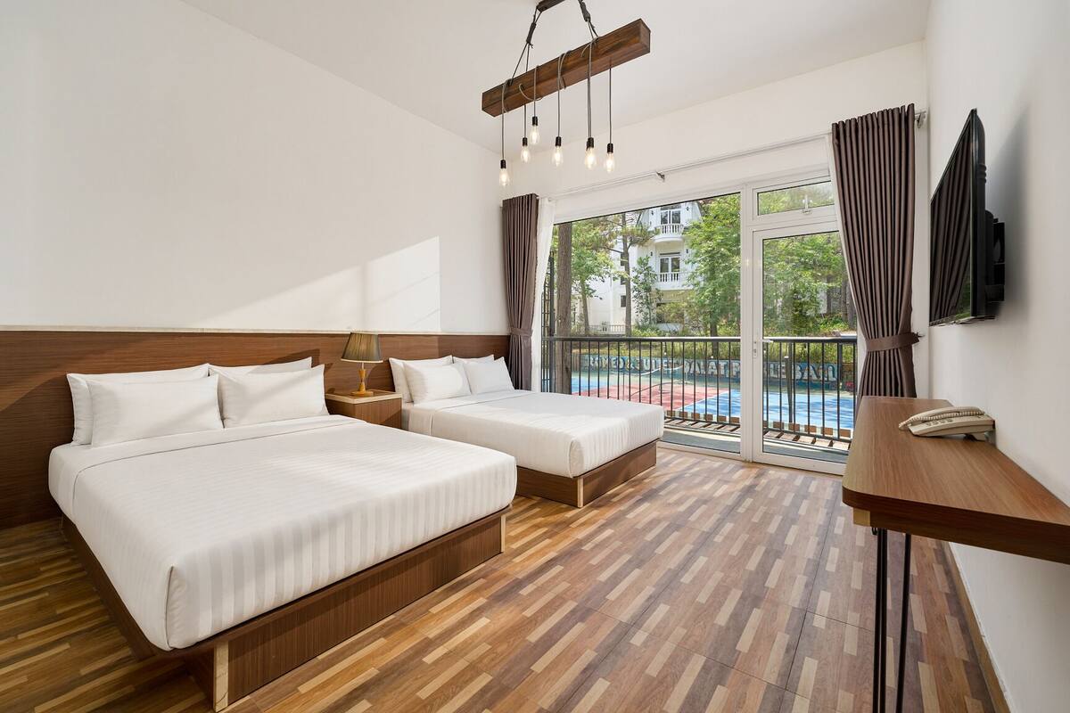 A hotel room with two beds, a wooden floor, a balcony with a view, and a TV on the wall.