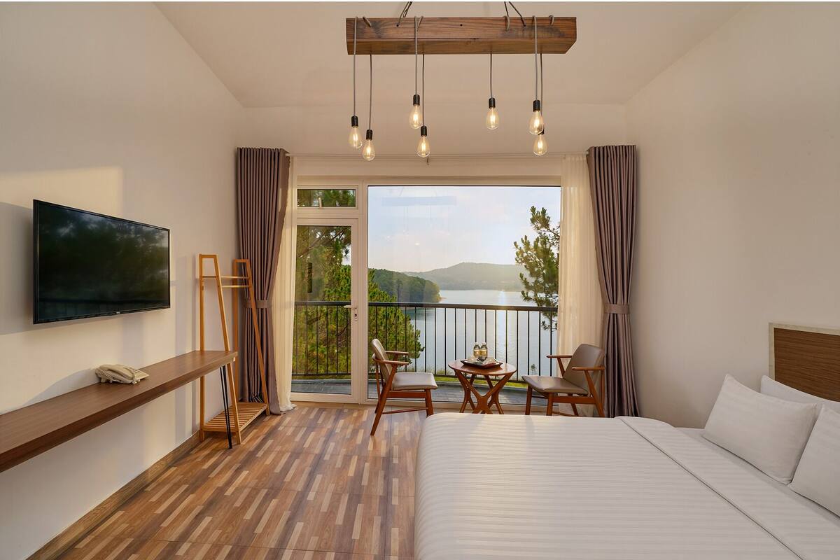 A hotel room with a bed, a TV, a balcony with a table and chairs, and a view of a lake.