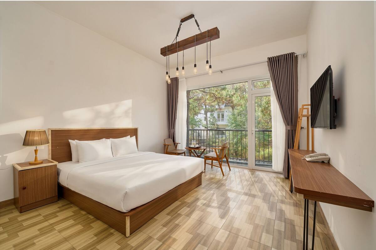 A hotel room with a large bed, a wooden nightstand, a desk, a chair, a table, a TV, and a balcony with a view.