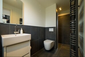 Un baño moderno con un lavamanos blanco, paredes de azulejos oscuros, un inodoro blanco y un perchero.