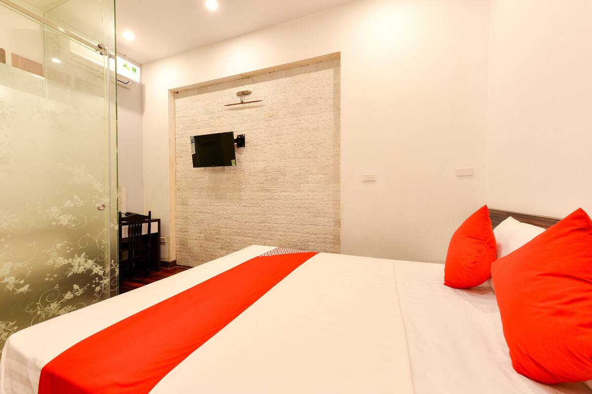Hotelkamer met een bed, rode kussens, een witte dekbedovertrek, een glazen douchecabine en een houten stoel.