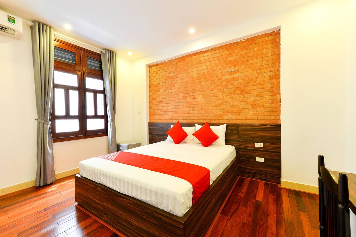Een hotelkamer met een houten bed, rode kussens, een accentmuur van baksteen en een raam met gordijnen.