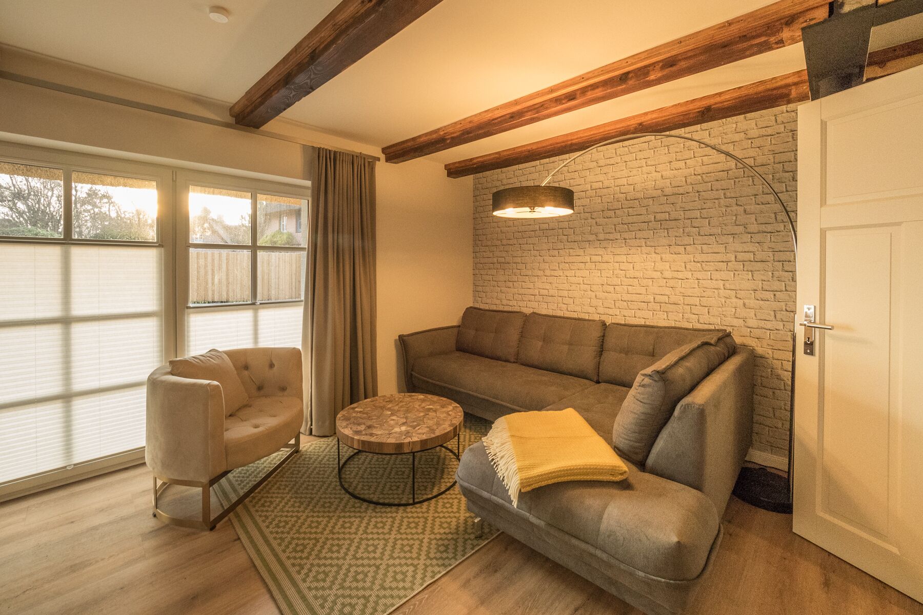 A cozy living room with a sofa, armchair, and coffee table, a brick wall, and a large window with blinds.
