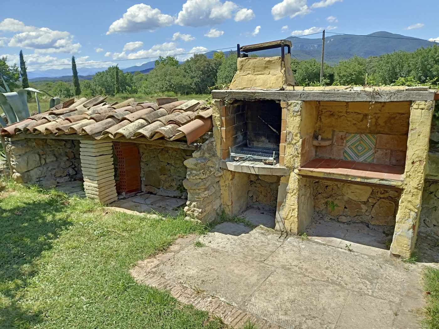 Un bâtiment rustique en pierre, avec un toit de tuiles et une cheminée, entouré de verdure et de montagnes.